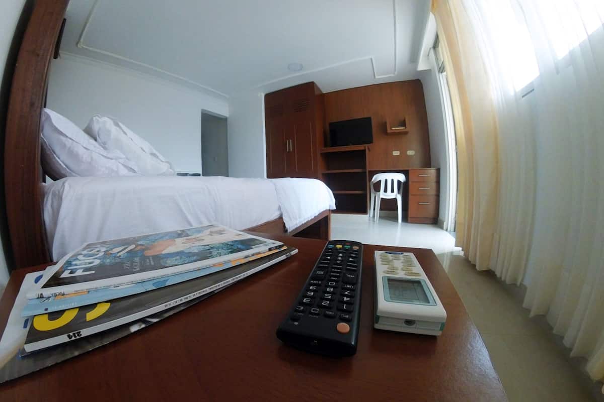 A hotel room with a bed, a desk with a remote control and a magazine, a TV, and a chair.