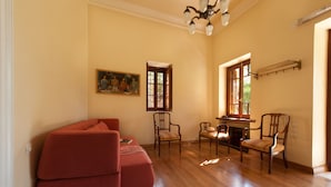 A spacious living room with hardwood flooring, a red sofa, two wooden chairs, a dining table, and a painting on the wall.