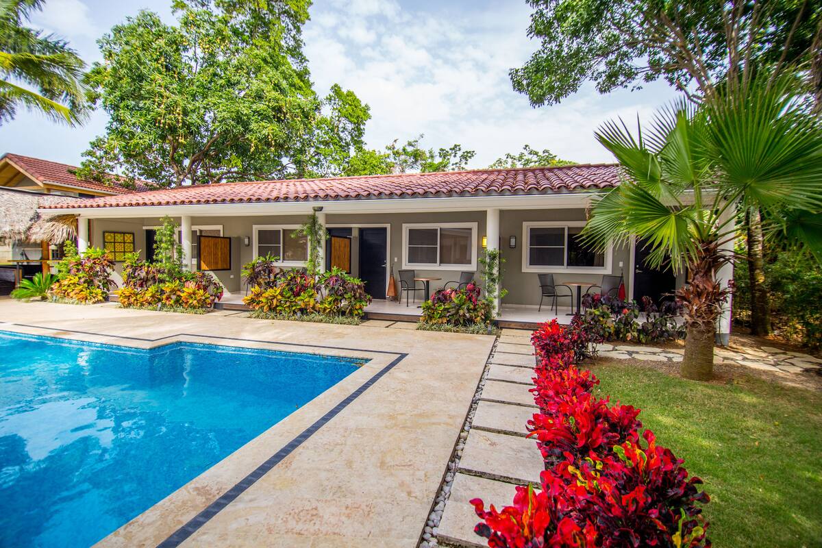 A two-story house with a swimming pool, surrounded by lush greenery and various plants.