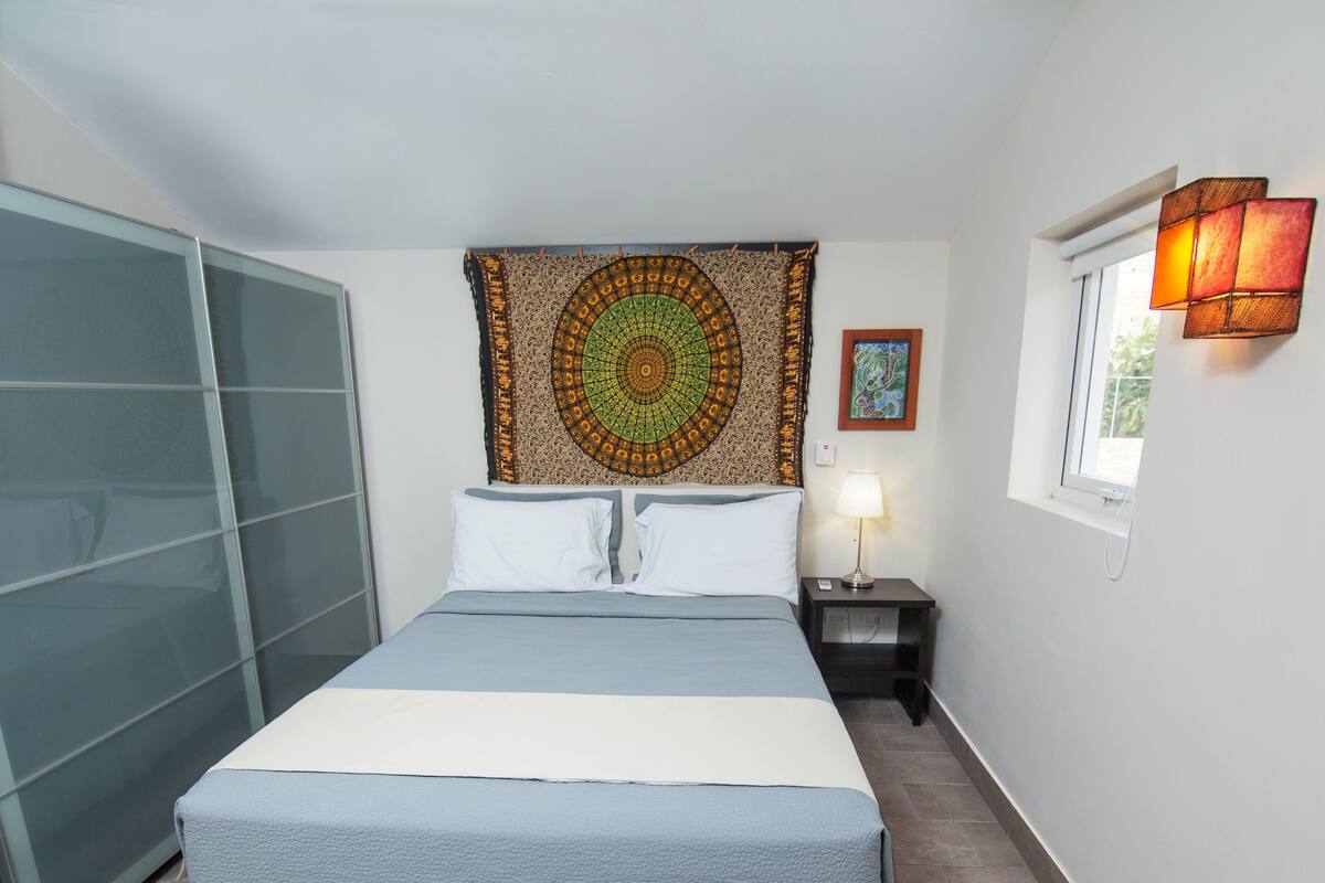 A bedroom with a bed, a nightstand with a lamp, a tapestry, and a window with a view.