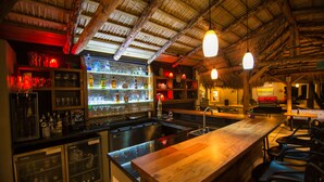 Uma área de bar com balcão de madeira, prateleiras com garrafas e teto de palha.