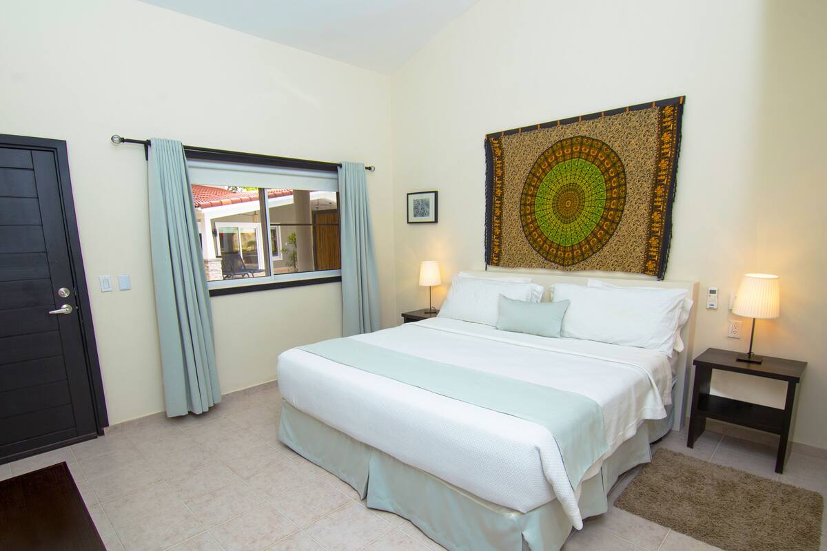 A hotel room with a bed, a window with curtains, a tapestry on the wall, and a nightstand with a lamp.
