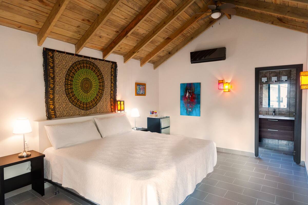 A bedroom with a large bed, wooden ceiling beams, a tapestry, wall art, and a bathroom visible through an open door.