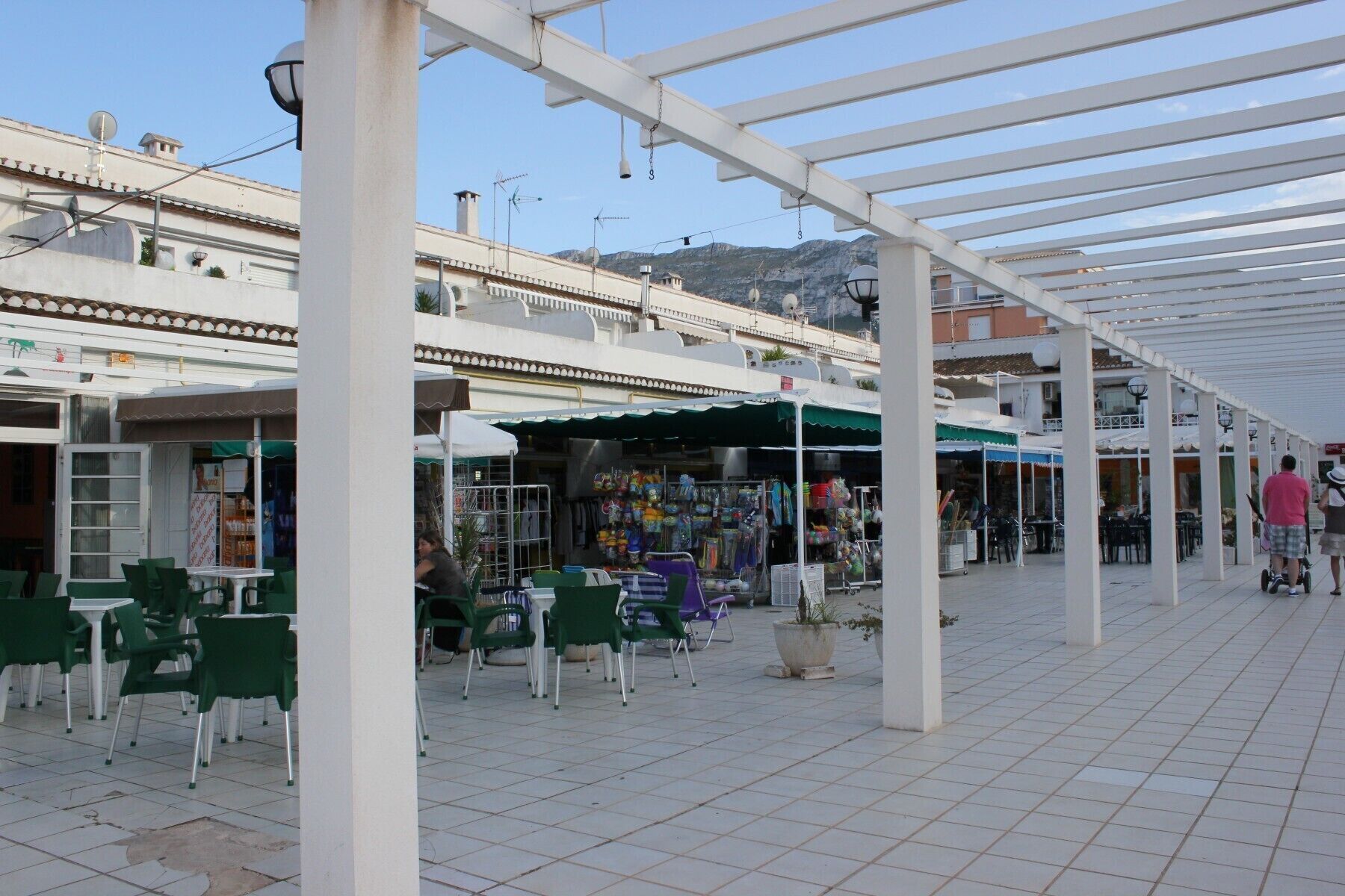 Un área de café al aire libre con sillas y mesas verdes, un puesto cubierto en un mercado que vende diversos artículos, y una estructura de pérgola blanca.
