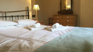 Een netjes opgemaakt bed met witte lakens en opgerolde handdoeken, een houten dressoir met spiegel, en een lamp op een nachtkastje.