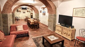 Een gezellige woonkamer met een bank, een salontafel en een televisie op een kast. Er zijn bogen en stenen muren, en een zithoek met stoelen en een tafel.