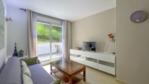 Una sala moderna con sofá, una mesa de centro de vidrio, un televisor sobre un mueble blanco y un balcón con vistas a zonas verdes.