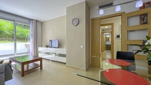 Una sala moderna con una mesa de centro de vidrio, un televisor en un mueble blanco, un reloj en la pared y vistas al exterior con vegetación.