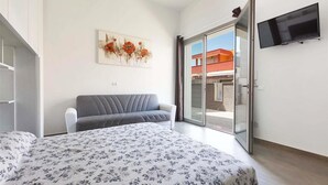 Un dormitorio con cama, sofá gris, un cuadro, televisor y puerta corrediza de vidrio hacia el exterior.