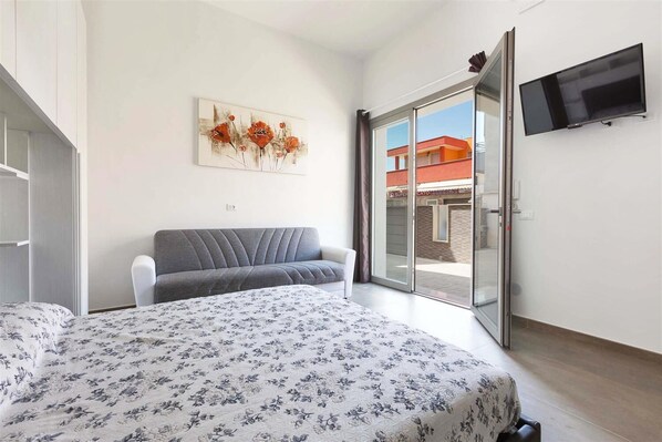 A bedroom with a bed, a grey sofa, a painting, a TV, and a sliding glass door to the outside.