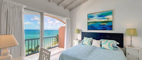 A bedroom with a bed, a painting, a nightstand, a lamp, and a view of the ocean.