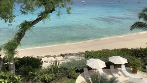 Blick auf einen Strand mit klarem türkisblauem Wasser, Booten und Liegestühlen mit Sonnenschirmen.