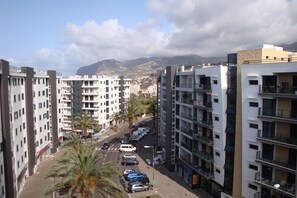 Un edificio residencial alto con varios balcones, un estacionamiento con coches y una calle bordeada de palmeras.