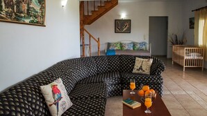 A living room with a black patterned sofa, a wooden staircase, a bed with floral pillows, a wooden crib, and a table with a book and glasses of orange juice. - Peaceful and private family villa, big pool, wellness, views of sea and mountain (Chania Crete)