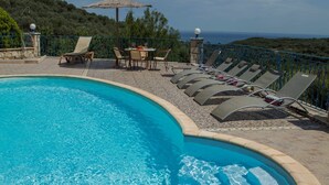 A pool area with lounge chairs and a table, surrounded by a railing and a view of the sea. - Peaceful and private family villa, big pool, wellness, views of sea and mountain (Chania Crete)