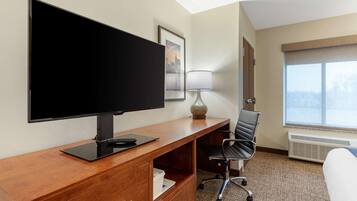 Habitación de hotel con una amplia televisión de pantalla plana sobre un escritorio de madera, una silla de oficina negra, una lámpara, una cama y una ventana con vistas.