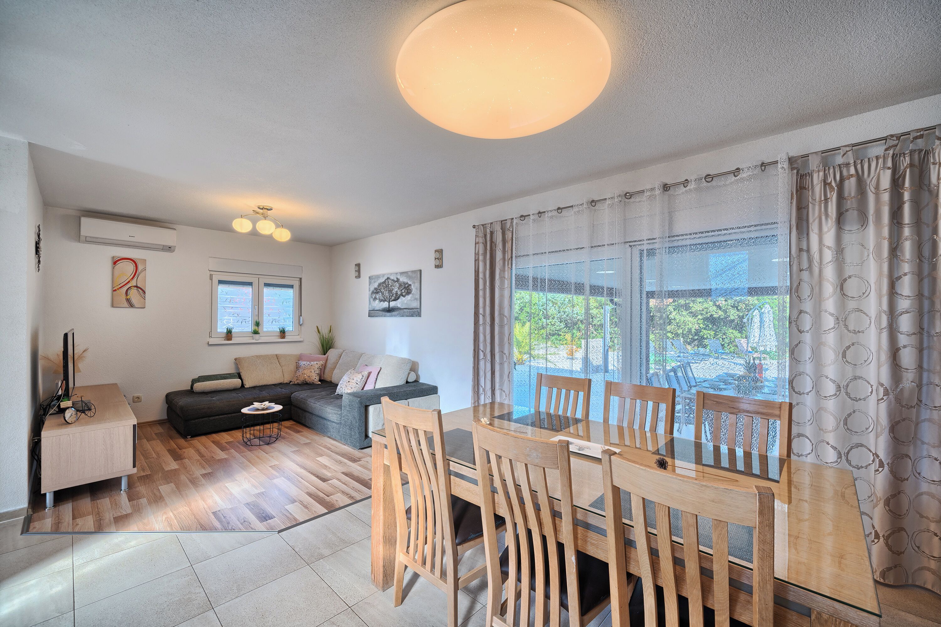 Uma sala de estar moderna com sofá, mesa de jantar com cadeiras, uma janela grande com cortinas e uma luminária no teto.