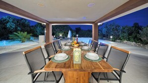 An outdoor dining area with a wooden table set for six, surrounded by chairs, under a covered patio with a view of a swimming pool and trees at dusk. - Luxury villa with heated pool, Marina-Trogir (Pozorac, Marina)