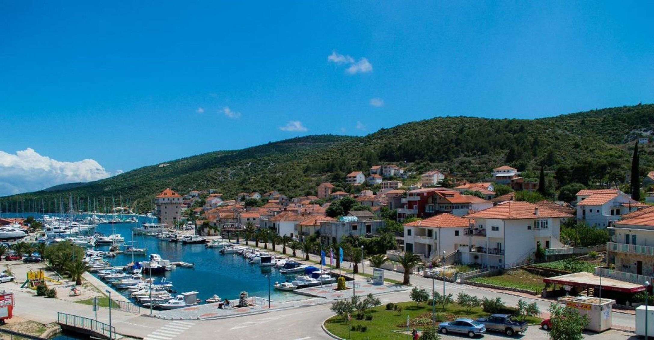 A marina with boats, a coastal town with red-roofed buildings, and a hilly backdrop.