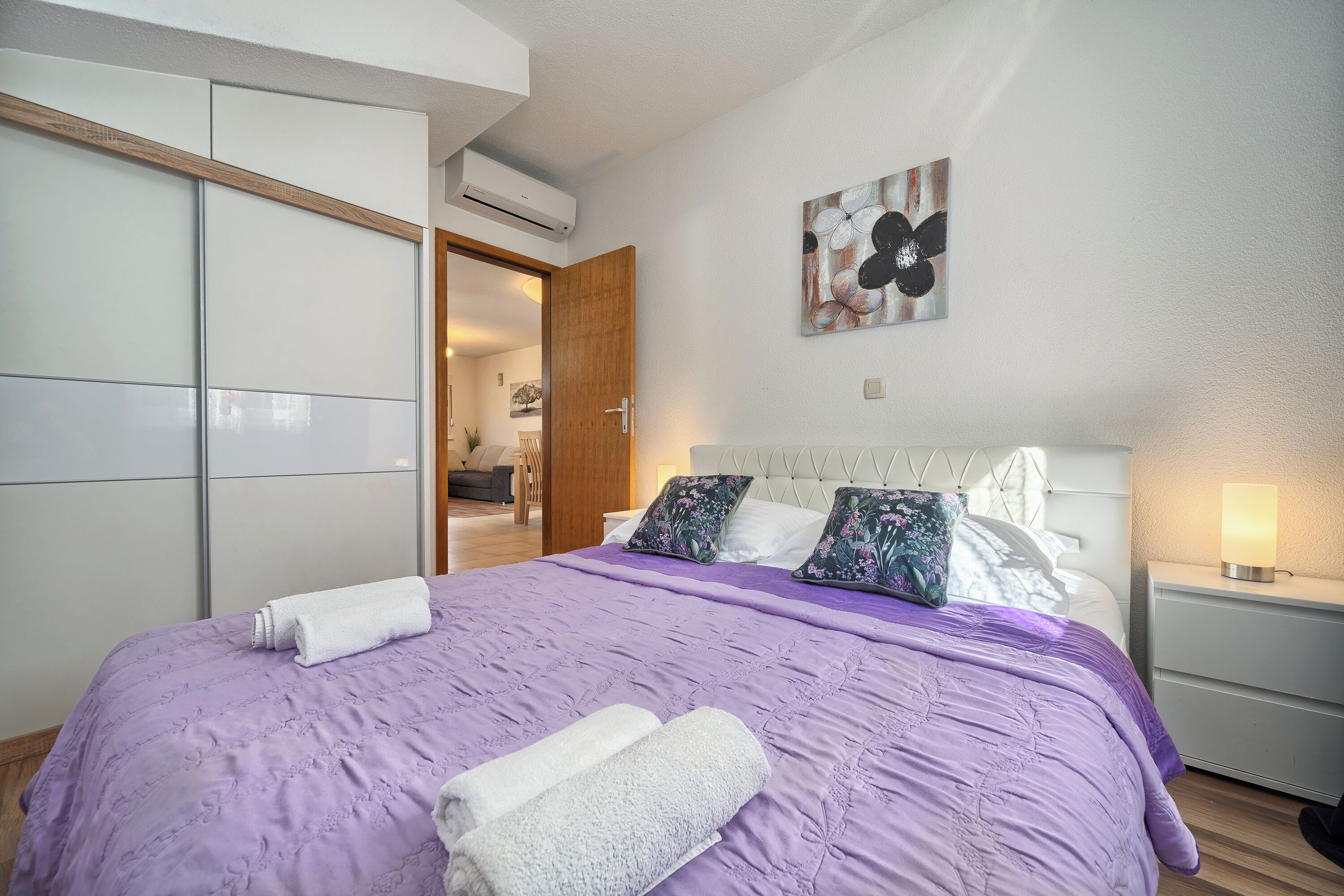 Une chambre à coucher comprenant un lit, une table de chevet, un climatiseur fixé au mur et une porte donnant sur une autre pièce.