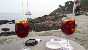 Dos vasos de bebida roja con fruta sobre una mesa frente al mar, con terreno rocoso y un velero al fondo.