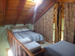 A room with a sloped ceiling, a bed with a striped bedspread, a bedside table, and a window with curtains. - Casita Pacifica- great 2BR/1BA house steps from Samara & Carrillo Beaches (Samara)
