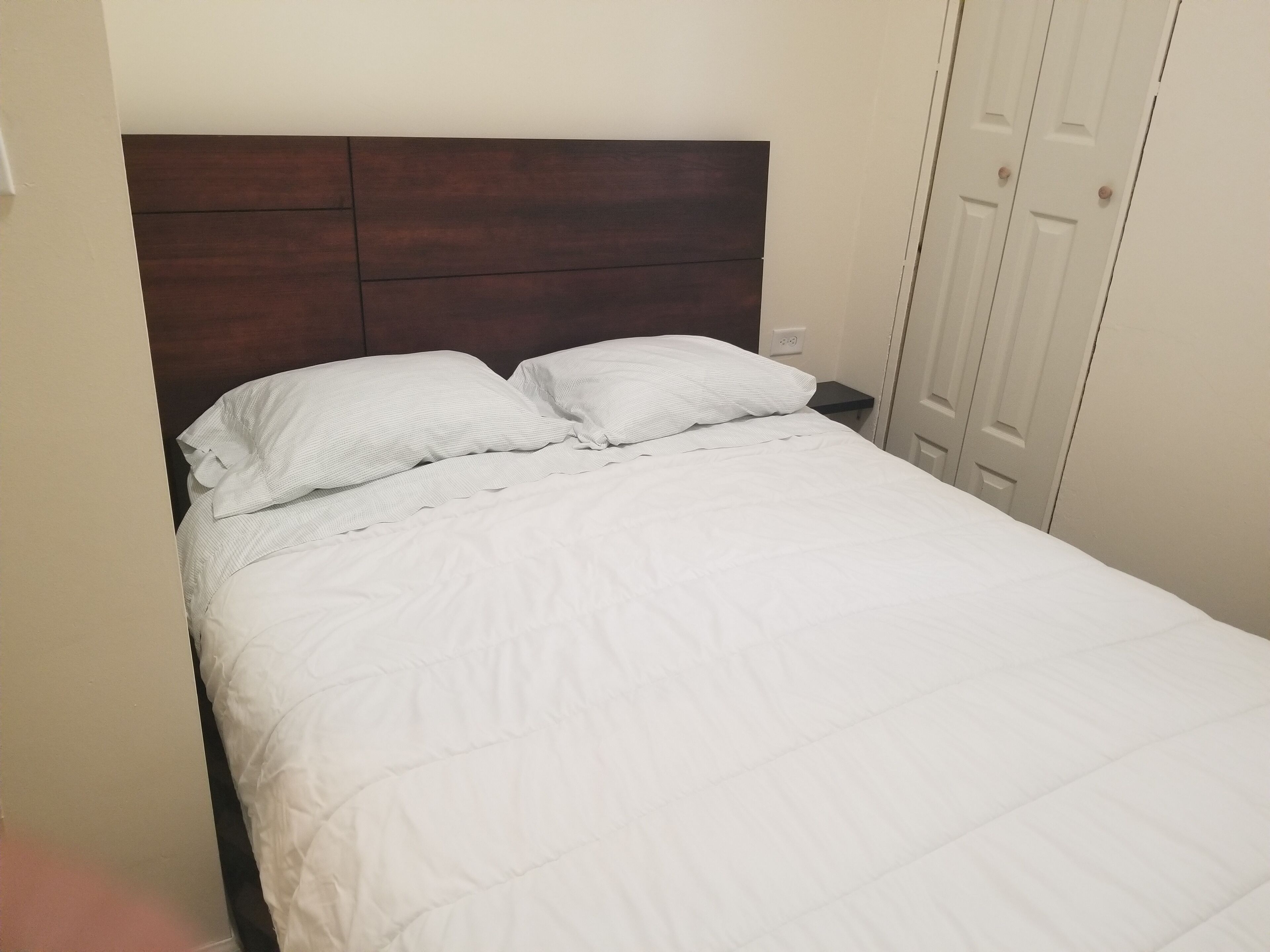 Ein ordentlich bezogenes Bett mit weißer Bettwäsche und zwei Kissen, ein dunkles Holz-Kopfende und eine weiße Tür im Hintergrund.