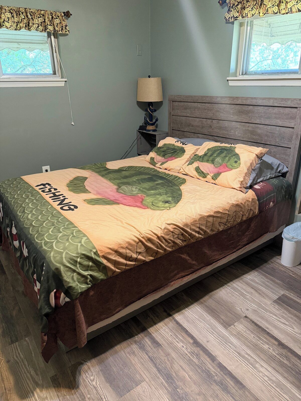 A bed with a green and pink fish-themed bedspread, a wooden headboard, a bedside lamp, and a window with a curtain.