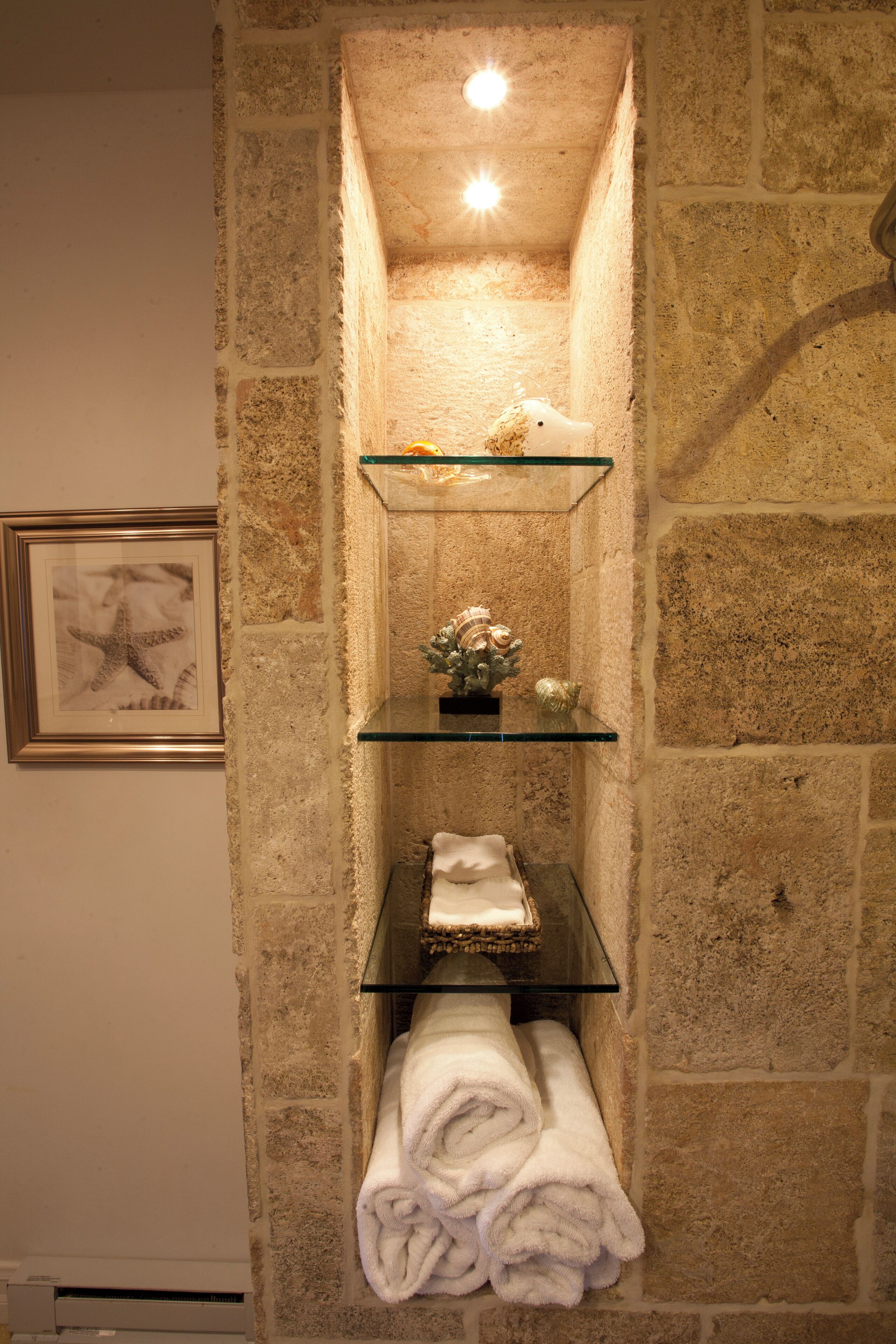 A bathroom with stone walls, a framed picture, and shelves with towels and decorative items.