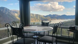 Ein Essbereich mit einem runden Tisch und Stühlen, großen Fenstern mit Blick auf Berge und See, sowie einem kleinen Tisch mit einer Kaffeemaschine.
