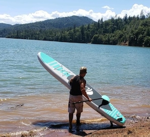 A person carrying a paddleboard near a lake with mountains in the background.