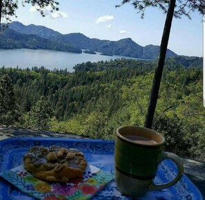 A scenic view of a lake surrounded by mountains and dense forest, with a table featuring a plate of food and a mug. - Secluded Romantic Getaway Overlooking Shasta Lake (Lakehead)