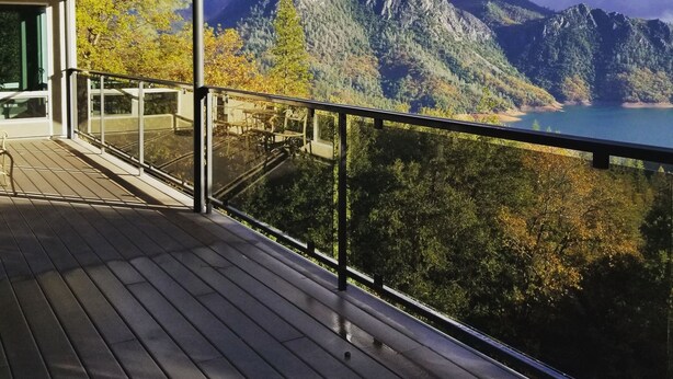 Eine Holzterrasse mit Glasgeländer, die auf eine Berglandschaft und ein Gewässer blickt.