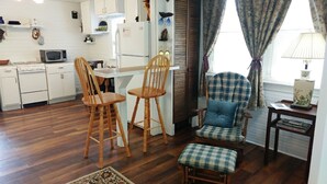 A compact kitchen with white cabinets, a microwave, and a stove. There are two wooden chairs and a blue plaid armchair. A small table with a lamp and a vase is near the window. - River's Edge Cabin - Swim - Kayak - Fish From Your Backyard !!!! (Franklin)