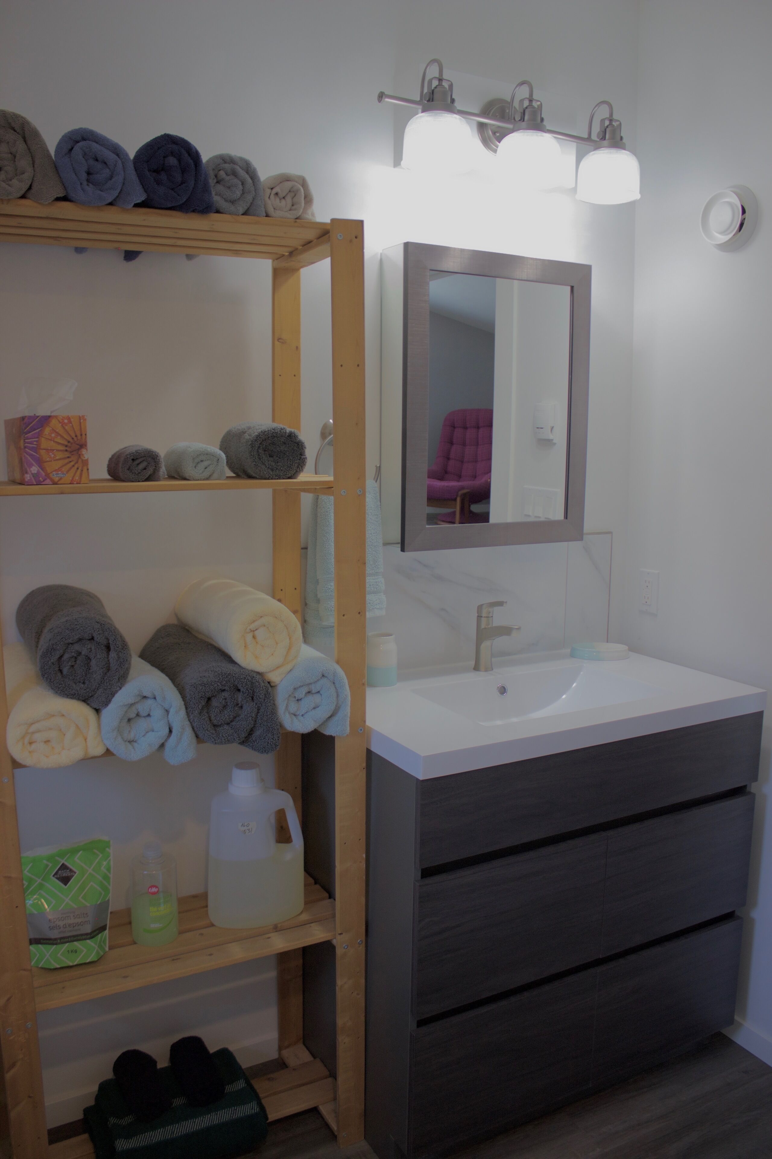 Une salle de bain avec une étagère en bois où sont pliées des serviettes, un lavabo blanc et un miroir au-dessus.