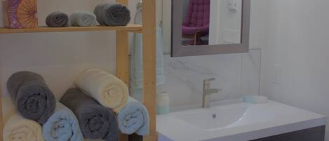 Une salle de bain avec une étagère en bois où sont pliées des serviettes, un lavabo blanc et un miroir au-dessus.