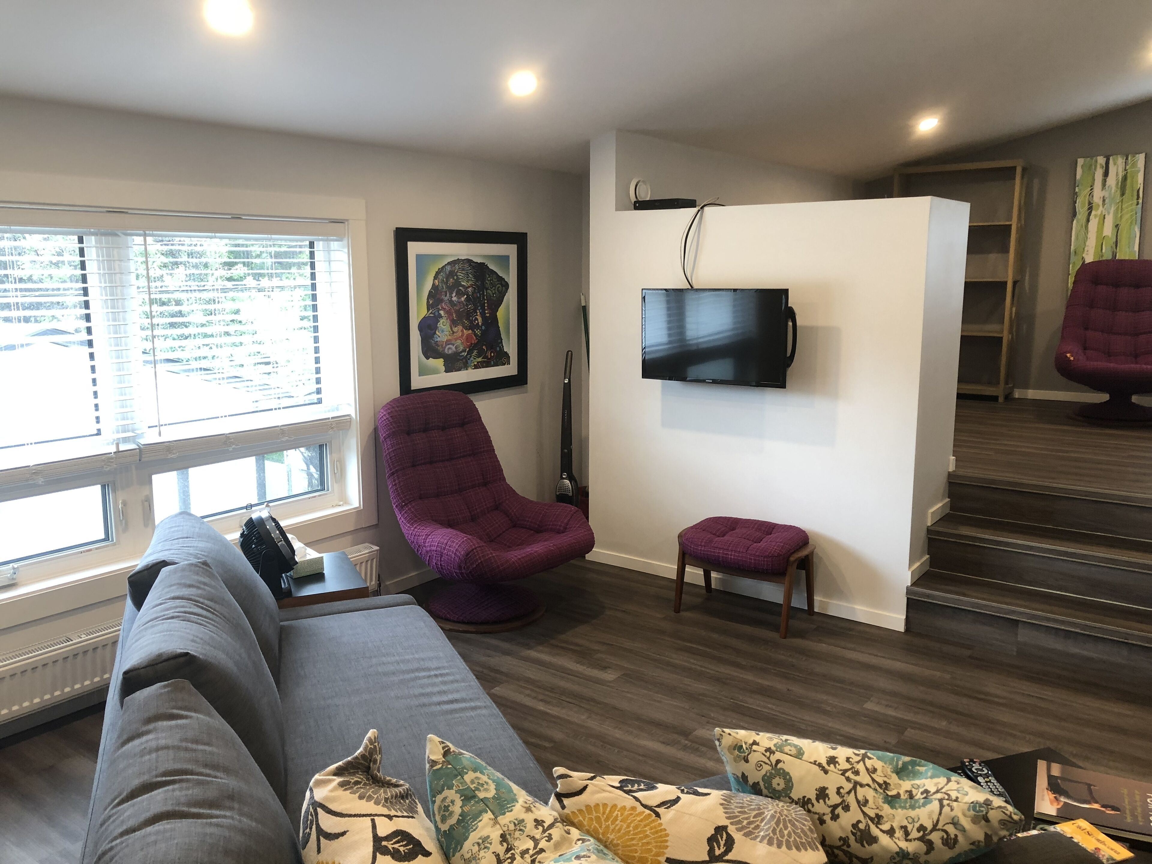 A modern living room with a grey sofa, a purple armchair, and a small ottoman. A flat-screen TV is mounted on the wall, and a framed artwork is displayed above it.