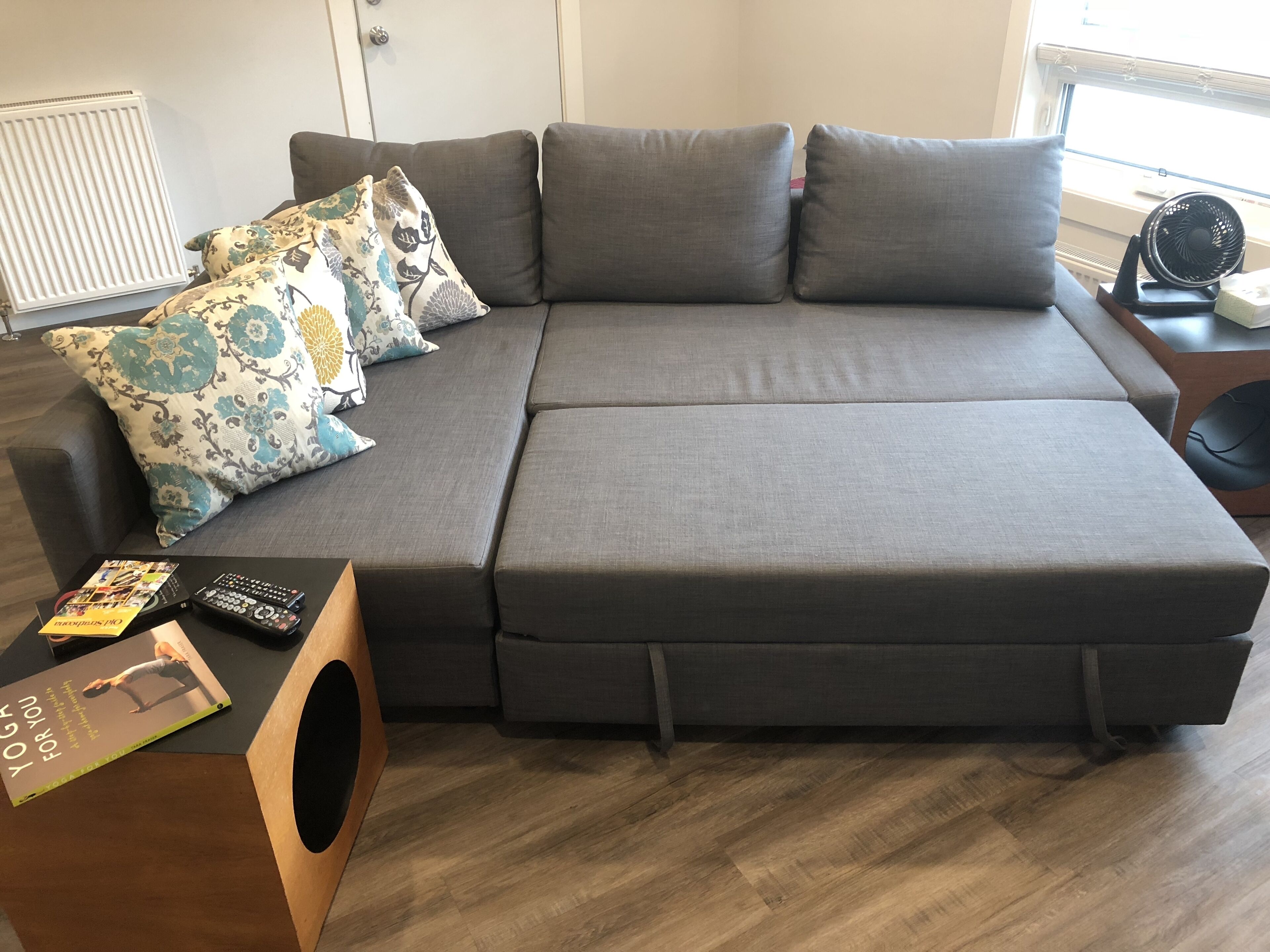 A grey sectional sofa with floral and plain cushions, a wooden side table with a book and remote controls, and a window with blinds.