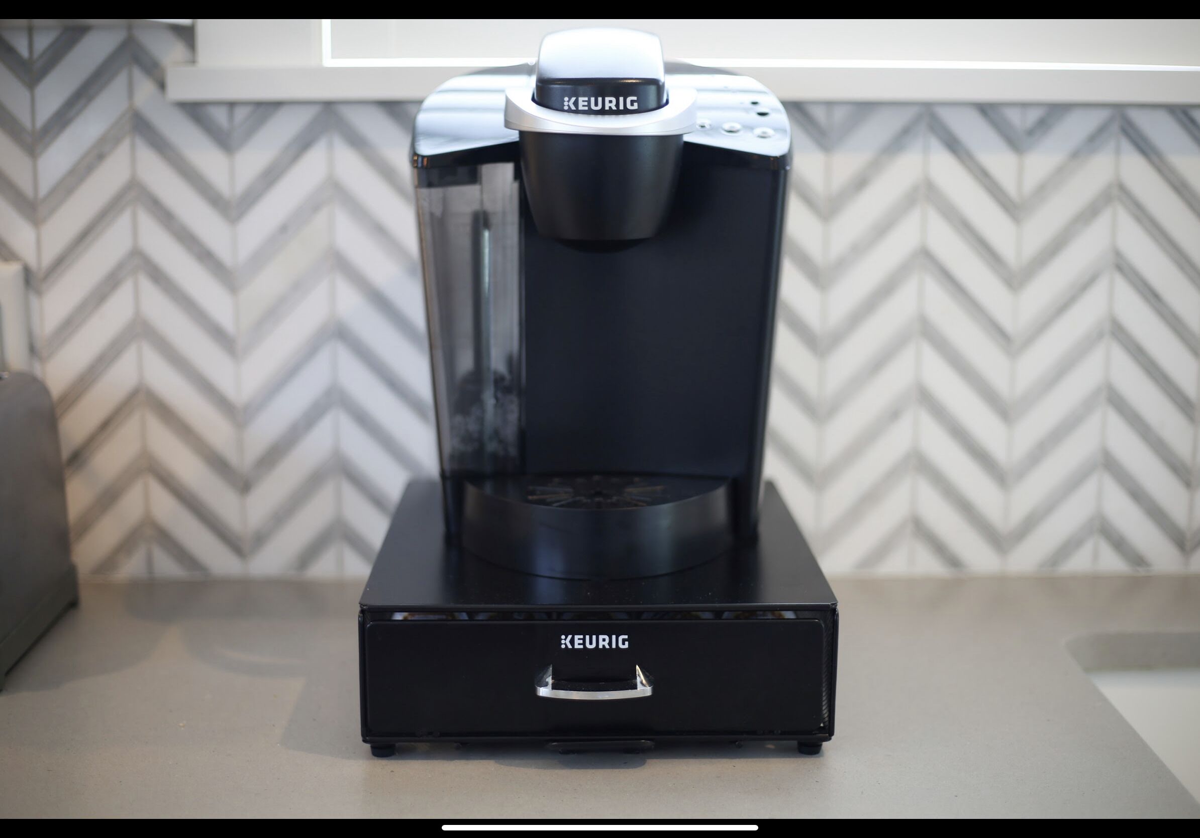 A Keurig coffee maker on a countertop with a chevron-patterned wall in the background.