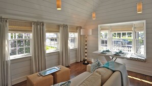 Ein Zimmer mit Holzboden, großen Fenstern mit Vorhängen, einer Couch, einem Tisch mit Pflanzen und einer Lampe.