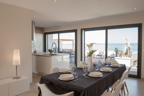 A dining area with a table set for four, large windows with a view of the sea, and a modern kitchen in the background.
