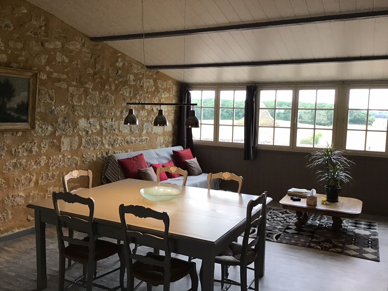 A dining area with a stone wall, a wooden table, and four chairs. There is a sofa with cushions in the background, and a window with a view of greenery.