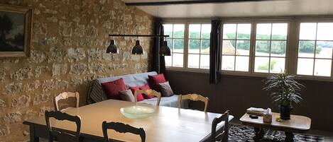 A dining area with a stone wall, a wooden table, and four chairs. There is a sofa with cushions in the background, and a window with a view of greenery.
