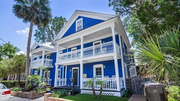 A two-story house with a blue exterior and white trim, featuring a front porch with white railings and a small front yard with plants and a wooden deck.