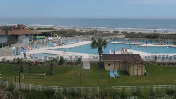 A beachside resort with a swimming pool, lounge chairs, and a small building, likely a lifeguard station.