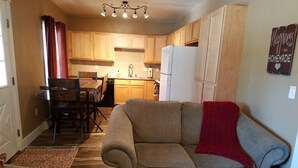 A cozy living room with a sofa and a small ottoman, wooden flooring, and a kitchen area with wooden cabinets and a refrigerator. - SADDLE MOUNTAIN ESCAPE (Clancy)