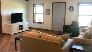 Una sala de estar con un televisor sobre un mueble blanco, una silla negra, un sofá color beige y una mesita.