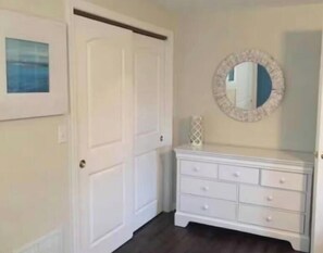 A room with a white door, a dresser with drawers, a round mirror with a decorative frame, and a framed picture on the wall.