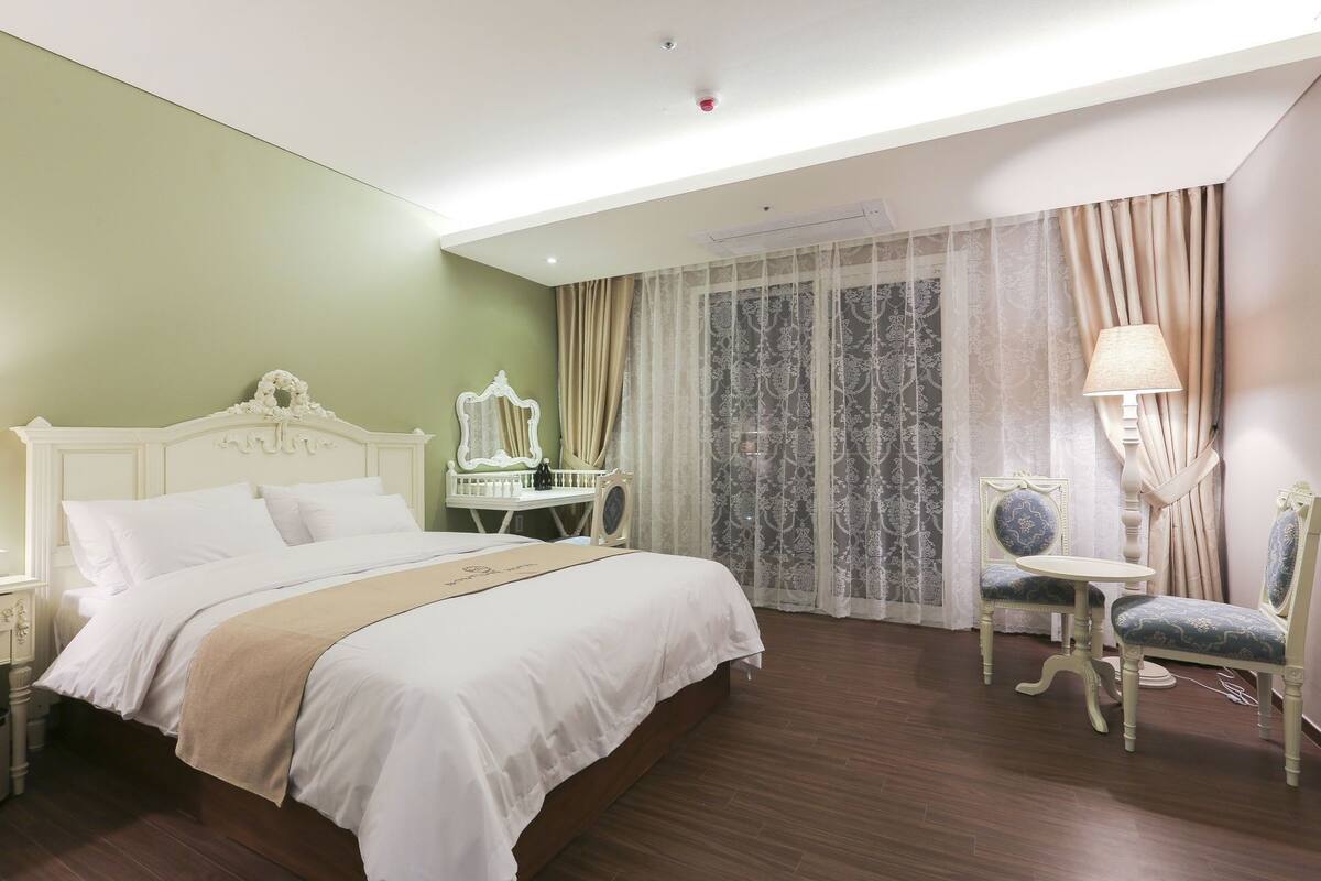 Habitación de hotel con cama, escritorio, silla, lámpara y ventana con cortinas.