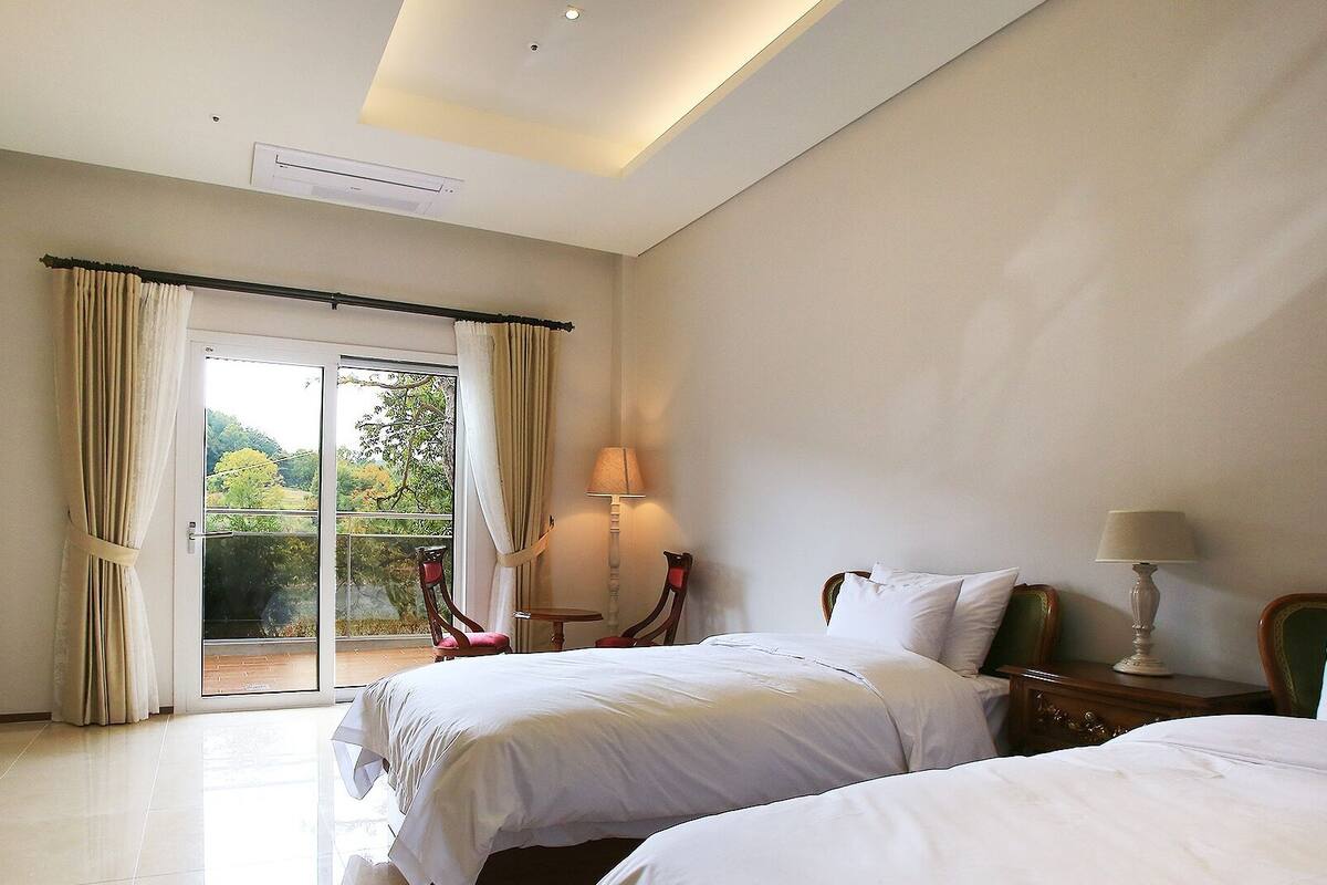Un dormitorio con dos camas, un ventanal con cortinas, un cuadro en la pared y una lámpara en una mesita de noche.